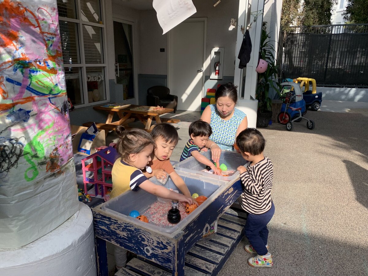 Kangaroo Room children exploring the outdoor sensory water table with their teacher